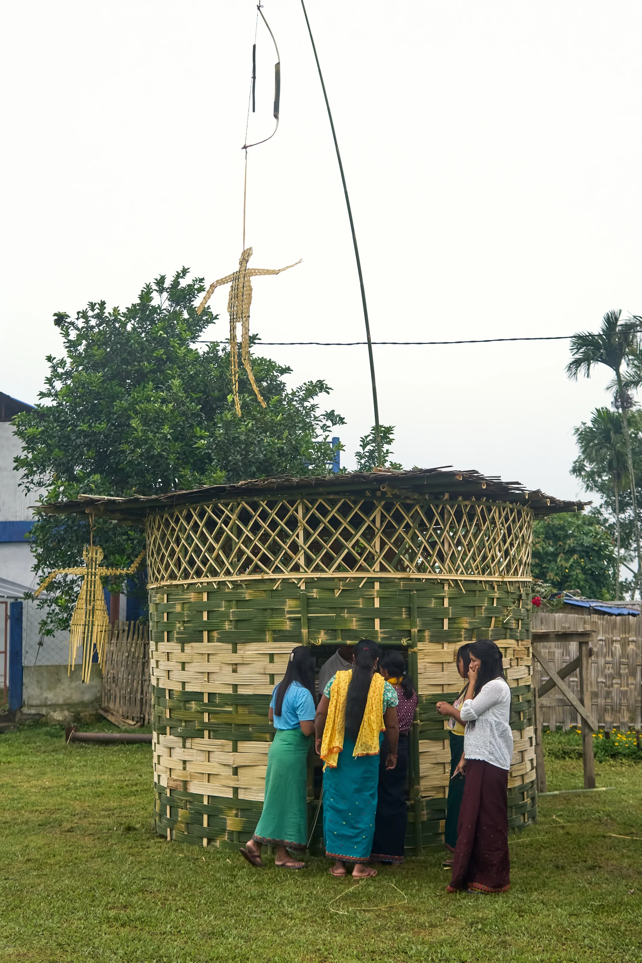 A woven bamboo installation with straw effigies hanging from its roofline, by day